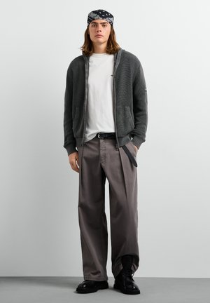 Giovane uomo che indossa una bandana nera, un cardigan grigio con zip, una t-shirt bianca, pantaloni larghi grigi, cintura nera e scarpe nere, in piedi davanti a uno sfondo semplice.