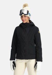 Schwarze isolierte Skijacke mit Reißverschluss, Kapuze und strukturierten Einsätzen. Weiße Handschuhe setzen einen Kontrast zu dem Outfit. Eine Strickmütze und eine Skibrille runden den Look ab.