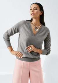 Grijze v-hals trui met geribde mouwen, gecombineerd met roze broek met wijde pijpen. Zilveren ketting met een ronde accent en een ring om de hand.