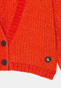 Cardigan lavorato a maglia arancione con finitura testurizzata, dotato di bottoni neri e una toppa circolare con il logo "cK" in bianco e nero.