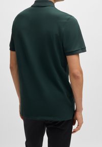 Mørkegrøn polo t-shirt lavet af glat stof, med klassisk krave og korte ærmer med ribbede manchetter. Synlige sideslids ved nederste kant.