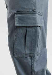 Gros plan sur la poche à rabat d'un pantalon cargo gris avec des coutures visibles sur le côté d'une jambe sur fond uni.