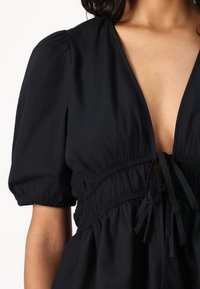 Blusa nera con scollatura a V profonda, maniche a sbuffo, vita arricciata e dettaglio con fiocco. Realizzata in tessuto liscio con una leggera lucentezza.