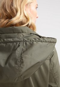 Olijfgroene capuchonjas met een gladde, gestructureerde stof. Heeft een brede kraag en gestructureerde schouders. Subtiele stikseldetails.