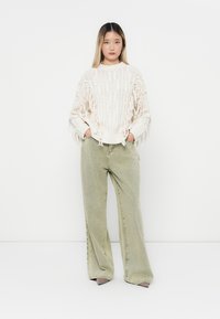 Elfenbeinfarbener Strickpullover mit Fransen, kombiniert mit olivgrünen weiten Jeans. Weiche Textur, lockere Passform und subtile Handwerkskunst.