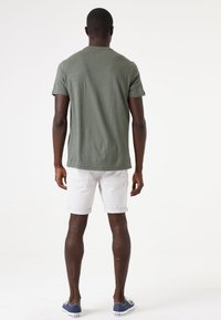 Olivgrünes T-Shirt mit kurzen Ärmeln, aus Baumwolle, klassische Passform, kombiniert mit weißen Shorts mit umgeschlagenem Saum. Hintergrund ist neutral.
