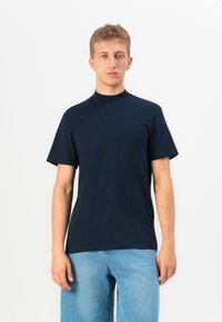 Giovane uomo in piedi, con una semplice maglietta nera a maniche corte e jeans azzurri chiari su uno sfondo bianco.