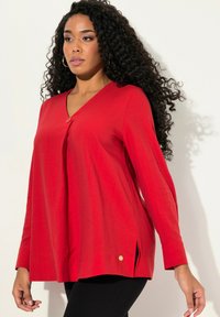 Top rosso a scollo a V con maniche lunghe, caratterizzato da dettagli con bottoni e spacchi laterali. Realizzato in un tessuto morbido e leggero con una texture liscia.