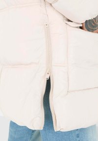 Pufferjack in lichtbeige, met een ritszak aan de zijkant en een gequiltde textuur. Hieronder zijn casual blauwe denim zichtbaar.