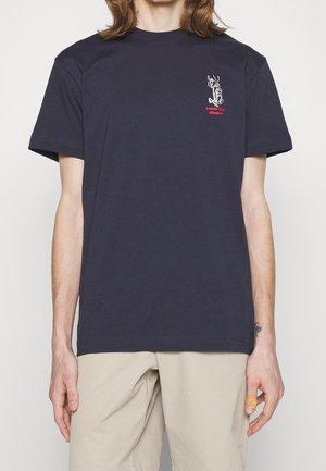 Person trägt ein marineblaues T-Shirt mit kleinem weißem Hummer und rotem "Lobster Club Athletics"-Text auf der oberen linken Brust, beige Hose und einen Verband am Finger.