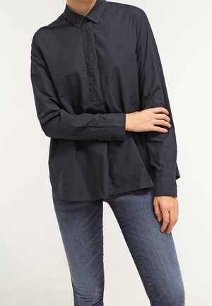 Donna che indossa una camicia nera larga con bottoni e maniche lunghe, e jeans slim fit blu, in piedi con un braccio incrociato sul corpo.