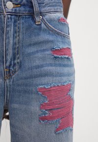 Oblečené modré džíny mají velké růžové nášivky na stehnech, roztrhané okraje a klasický design se pěti kapsami a zipem.