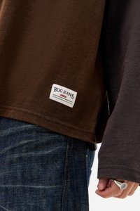 Κοντινό πλάνο ατόμου που φοράει σκούρο καφέ μακρυμάνικο πουκάμισο με ετικέτα BDG Jeans και μπλε τζιν παντελόνι, δείχνοντας το χέρι με ασημένιο δαχτυλίδι.