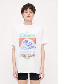 Valkoinen ylisuuri t-paita, jossa on värikäs graafinen kuvio aaltoa ja teksti "Surf Camp". Lyhyet hihat ja pyöreä kaula-aukko.