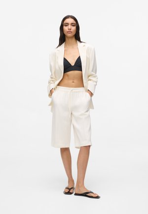 Femme portant un blazer crème, un short long assorti, un bralette noir et des tongs noires, debout les mains dans les poches sur fond blanc.