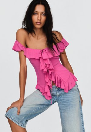 Woman wearing off-shoulder ruffled pink top and light blue denim shorts, posing with one leg bent against a plain background.