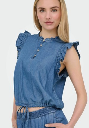 Młoda kobieta w niebieskim bezrękawniku z denimu z falbankami na krawędziach i dopasowanych spodniach ze ściągaczem w talii na jednolitym tle.