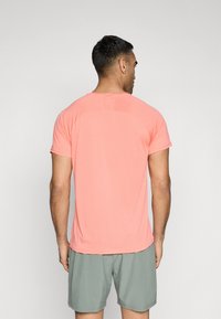 Tricou sport cu maneci scurte în nuanță de coral, având o textură ribbed și un gât rotund, asociat cu o pereche de pantaloni șort gri, prezentat din spate.
