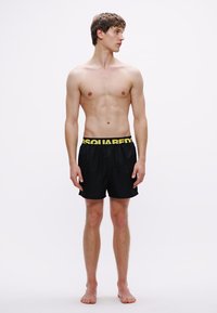 Fekete úszónadrág sárga, rugalmas derékpánttal, logóval díszítve. Az anyag könnyűnek és enyhén fényesnek tűnik. A modell felső nélkül van.