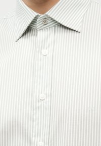 Κλείσιμο πλάνου ενός ατόμου που φοράει πουκάμισο με κάθετες λευκές και ανοιχτό γκρι ρίγες, με μυτερό γιακά και λευκά κουμπιά.