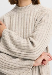 Suéter beige de canalé con un cuello alto y mangas anchas, fabricado en un tejido suave y texturizado. Líneas horizontales notables recorren la superficie.