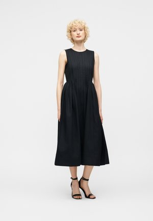 Žena s krátkymi kučeravými blond vlasmi, oblečená v čiernych midi šatách bez rukávov a čiernych sandáloch na vysokom podpätku so zapínaním okolo členka, stojaca pred jednofarebným pozadím.