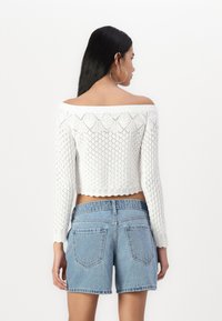 Fehér kötött, egy vállra húzható crop top, textúrált mintával. Világoskék farmer rövidnadrággal párosítva, hátul zsebekkel és rojtos szélekkel.
