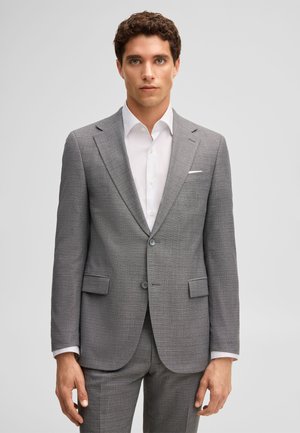 Man draagt een op maat gemaakt grijs pak met inkeping revers, wit overhemd en pochet, staand tegen een effen lichte achtergrond.