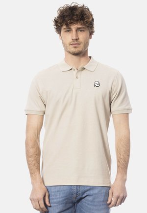 Man met krullend haar, gekleed in een beige poloshirt met korte mouwen en een klein logo op de borst, draagt een blauwe spijkerbroek en staat tegen een witte achtergrond.