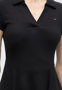 Gros plan d'une personne portant une robe noire côtelée à manches courtes avec un col et un petit logo Tommy Hilfiger rouge, blanc et bleu sur la poitrine.