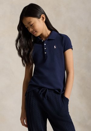 Κορίτσι με μακριά σκουρόχρωμα μαλλιά, που φοράει μπλε ναυτικό πόλο και υφασμένα μπλε ναυτικά παντελόνια, κοιτάζοντας προς τα κάτω με το ένα χέρι στην τσέπη.