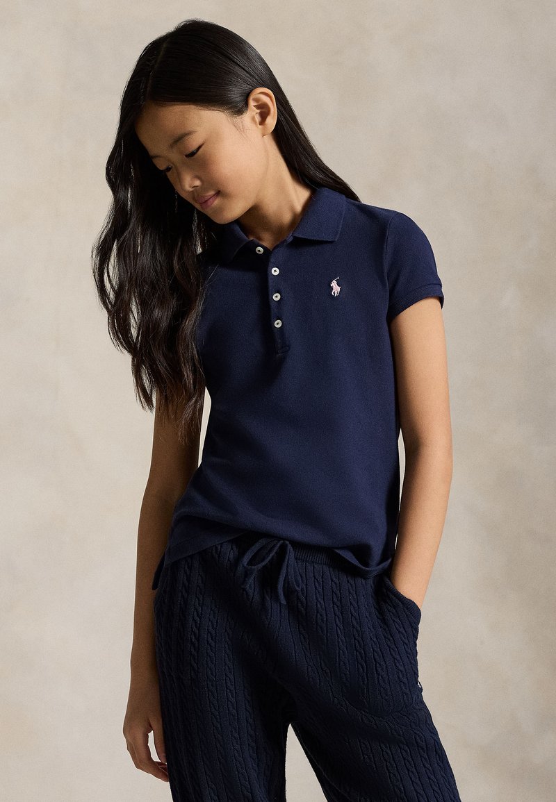 Mergina ilgais tamsiais plaukais, dėvinti tamsiai mėlyną polo marškinėlį ir tekstūruotas tamsiai mėlynas kelnes, žvelgia žemyn, viena ranka kišenėje.