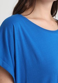 Modré bavlněné tričko s širokým výstřihem a krátkými rukávy, které má hladkou texturu a minimální šití.
