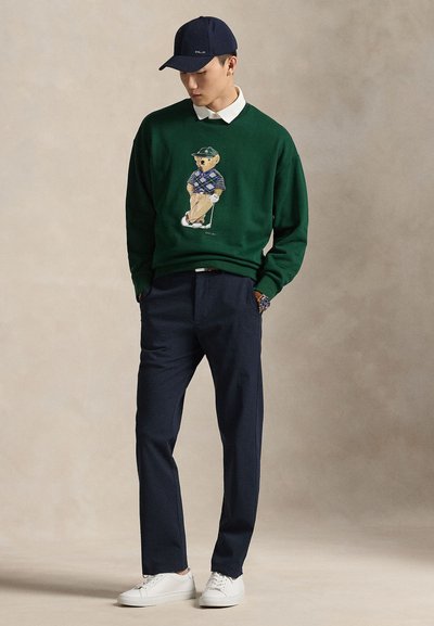Πράσινο πουλόβερ με κεντημένη αρκούδα σε γκολφ εμφάνιση, συνδυασμένο με ναυτικά μπλε παντελόνια και λευκά αθλητικά παπούτσια. Ένα ναυτικό μπλε καπέλο ολοκληρώνει την εμφάνιση.