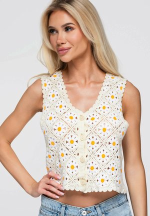Kvinde med langt blond hår iført en ærmeløs hvid hæklet top med gule og hvide blomster mønstre og knapper, kombineret med blå jeans.