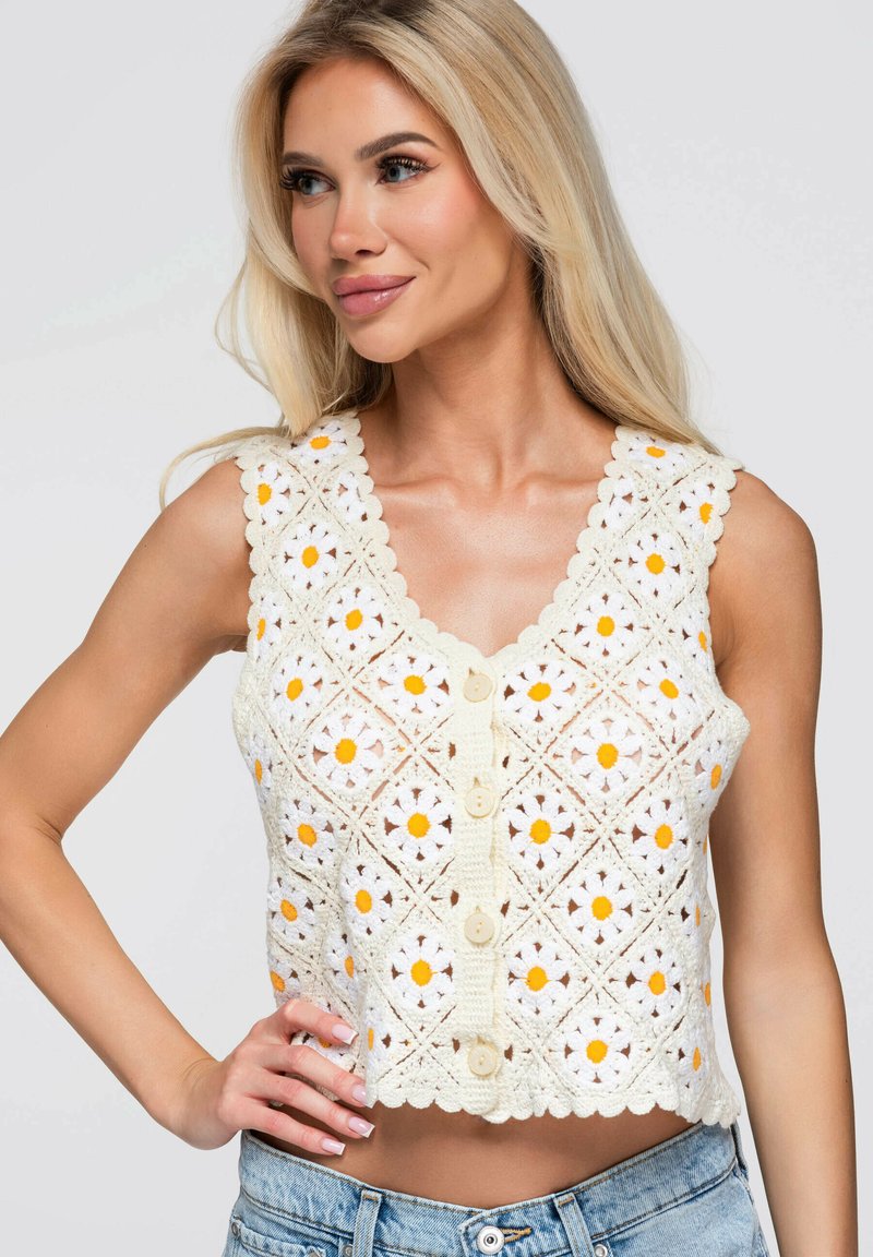 Femme aux longs cheveux blonds portant un haut blanc sans manches en crochet avec des motifs floraux jaunes et blancs et des boutons, associée à un jean bleu.