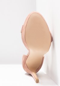 Chaussure à talon haut en suède rose blush pour femme, posée sur la semelle contre un mur blanc, montrant la semelle texturée et le talon fin.