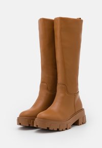 Wysokie brązowe skórzane botki z grubymi, bieżnikowanymi podeszwami i gładkimi cholewkami, stojące obok siebie na białym tle.