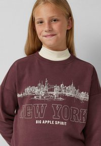 Felpa color bordeaux con una grafica bianca dello skyline di New York con il testo "NEW YORK" e "BIG APPLE SPIRIT" in caratteri in grassetto.