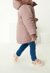 Lichtroze pufferjack met nepbont muts, blauwe jeans en pastelroze sneakers met bloemenaccenten en klittenband.