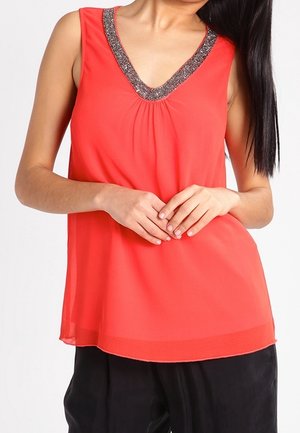 Kobieta ubrana w bezrękawową koralową bluzkę z metalicznym, koralikowym dekoltem w kształcie litery V, z rękami splecionymi, w połączeniu z luźnymi czarnymi spodniami.