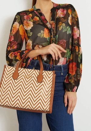Sac en toile de couleur crème avec un motif en zigzag marron, doté de poignées et d'accents en cuir marron. Présenté avec un chemisier fleuri et un jean.