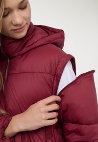 Gilet imbottito rosso con collo alto e chiusura con cerniera; presenta un esterno liscio e lucido e uno strato interno visibile sotto il braccio.