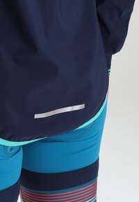 Coupe-vent marine avec accents turquoise et bande réfléchissante, associé à un short ajusté bleu sarcelle et marine avec motifs rayés et matériau texturé.