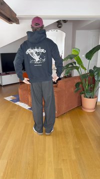 Μαύρη hoodie με λευκό γραφικό σχέδιο στην πλάτη, γκρίζες φόρμες, στέκεται σε ξύλινο δάπεδο σε δωμάτιο με φυτά και έπιπλα.
