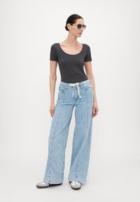 Grå tettsittende t-skjorte, lyseblå vide denimjeans med snor i livet og sidesømmer. Modellen har på seg mørke solbriller og uformelle joggesko.