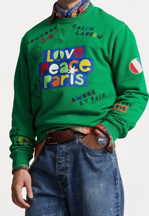Mies, jolla vihreä collegepaita, jossa värikäs teksti "Love Peace Paris" ja rauhanmerkkejä, ruudullinen paita alla, farkut, ruskea vyö ja rannekello.