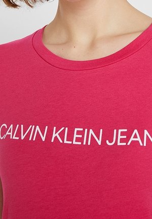 Woman wearing a bright pink crew neck t-shirt with "CALVIN KLEIN JEANS" printed across the chest in white letters.