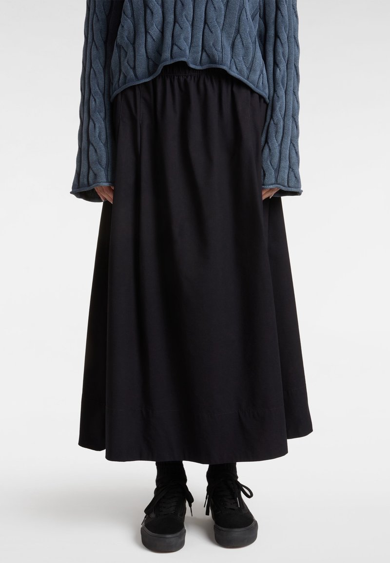 Personne portant une longue jupe noire, des baskets noires et un pull court bleu en maille torsadée, debout sur un fond blanc uni.