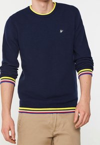 Marinblå tröja med strukturerad stickning, rund halsringning och kontrasterande gula, röda och blå randiga ärmar och nederkant. Litet logotyp på bröstet.